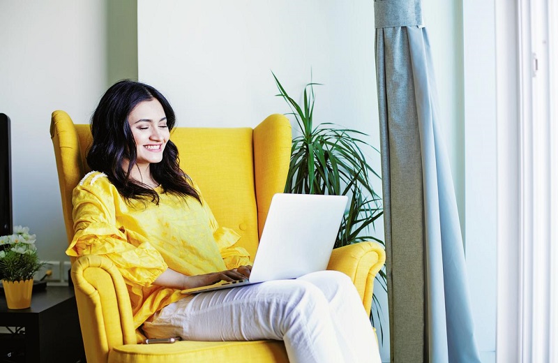 Lady sitting on a couch while smiling and working on a laptop
