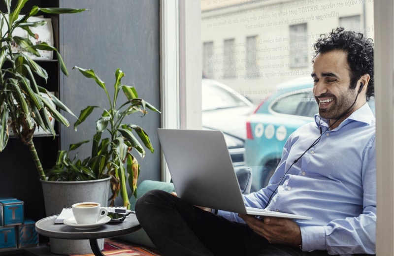 Man sitting on a seat by a window, smiling at a laptop sitting on his lap