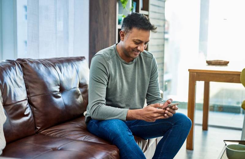 A man sitting on a couch smiling while using his phone